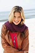 A young blonde woman on a beach wearing a brown leather jacket, a pink top and a woollen scarf