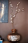 Branch of white flowers in ornate wooden vase and partially visible picture on brown wall