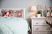Girl's bedroom - rag doll on embroidered pillow on bed with headboard next to table lamp and soft toys on white chest of drawers