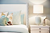 Various scatter cushions on bed with tall, white upholstered headboard next to table lamp with white ceramic base and fabric lampshade on bedside cabinet