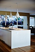 White kitchen island with integrated sink below group of pendant lamps with glossy, bottle-shaped lampshades in modern interior with floor-to-ceiling terrace doors