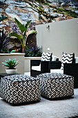 Patterned pouffes and black armchairs with patterned scatter cushions on terrace with tropical plants in raised beds against rock face and masonry wall