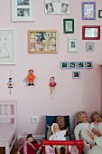 Puppen in Puppenwägen vor rosa getönter Wand, mit gerahmten Bildern und Hampelmänner