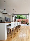 Bar stools with white loose covers at kitchen counter in open-plan designer kitchen; open folding terrace doors in background