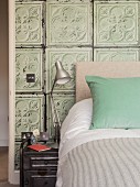 Table lamp on metal bedside cabinet next to bed with turquoise scatter cushion against wall covered in old 3D structured tiles