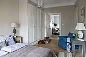 Classic bedroom in white and natural shades with blue accents; view over bed to fitted wardrobes and dining area in adjoining room