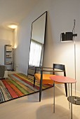 Filigraner Beistelltisch mit roter Platte und Armlehnstuhl mit orangem Polsterbezug vor Stehleuchte, daneben Standspiegel auf buntem Streifenteppich in minimalistischem Ambiente