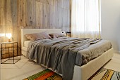 Elegant bedroom - double bed with upholstered frame and headboard against wood-clad wall and lit table lamp on delicate bedside table made of metal rods