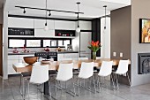 Dining table with 10 light shell chairs in front of modern, white fitted kitchen