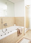 Fitted bathtub against half-height, sand-coloured tiles below large mirror on wall and separate shower cubicle with glass door in background