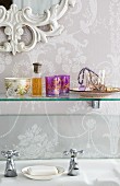 Ornate mirror frame, floral wallpaper and glass shelf of romantic knickknacks above vintage tap fittings
