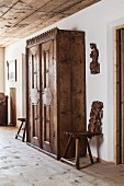 Ornate, antique wooden wardrobe flanked by plank chairs and carved effigy of saint on wall