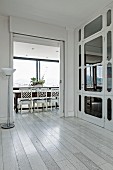 Foyer with white-stained wooden floor and standard lamp next to open doorway leading to dining area with traditional, white wooden chairs