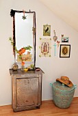 Branch of flowering quince in glass vase on antique metal cabinet in front of mirror with romantic decorations and next to gallery of pictures above African laundry basket