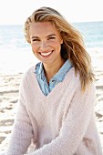 A young blonde woman by the sea wearing a denim shirt and a white woollen jumper