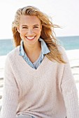 A young blonde woman by the sea wearing a denim shirt and a white woollen jumper