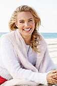 A young blonde woman by the sea wearing a light knitted jumper with another jumper over her shoulders