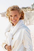 A young blonde woman on a beach wearing a white coat with a jumper over her shoulders