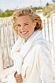 A young blonde woman on a beach wearing a white coat with a jumper over her shoulders