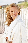 A young blonde woman on a beach wearing a white coat with a jumper over her shoulders