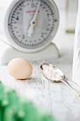 An egg and a spoonful of flour in front of a pair of kitchen scales