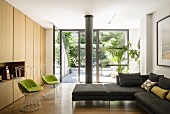Minimalistisches Wohnzimmer mit modernen Drahtstühlen, hellen Holzfronten und großer Glasfront mit Blick in den begrünten Innenhof