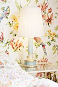 Wallpaper with pattern of flowers and parrots, table lamp with pleated lampshade on bedside table and partial view of bed