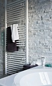Weisser Handtuchtrockner mit aufgehängten Handtüchern vor Wand mit weissen Verblender Riemchen, Naturstein Look, davor teilweise sichtbare Badewanne