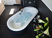 A relaxing looking, water-filled bathtub positioned at an angle in the room with modern taps and and overflow in a dark brown tone
