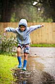 Junge in Gummistiefeln & Regenjacke springt in Pfütze herum