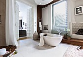 A free-standing, limestone bathtub and an artificial leather armchair in a white, cosy bathroom with built-in teak units and a view through a frame and panel door into the adjoining bedroom