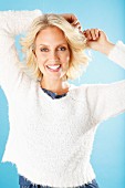 A young blonde woman with her hands above her head wearing a white knitted jumper