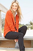 A young blonde woman sitting outside on a wooden bench wearing an orange cardigan