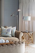 Tea break in elegant living room - tea service on tray on ottoman, sofa with elegant scatter cushions, matching table lamp on side table and stainless steel standard lamp