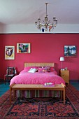 Chandelier above art nouveau bed and bedroom bench with cane seat; small painting on wall painted raspberry and pink bedspread