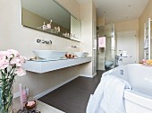 A wall console with a glass surface and two oval washing bowls with a custom-built mirror and a glass shelf above it and a shower cubicle in the background