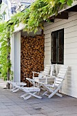 Seating area with white deckchairs and side table below wisteria growing over outside one-storey extension with stacked firewood