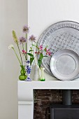 Pewter plates and collection of small vases containing a few flowers on mantelpiece