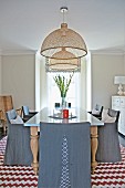 Elegant, grey, loose-covered chairs around dining table below two wicker pendant lamps and rug with red and white zigzag patterns in country-house interior with French windows