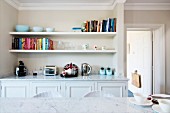 View across marble table top to fitted sideboard in niche below books and ornaments on two white, floating shelves in vintage ambiance