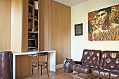 Brown leather easy chairs below artwork, wooden fitted cupboards with integrated desk and Thonet chair