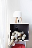 Garland of lanterns in various shades of grey in shelf compartment; table lamp with white lampshade on top