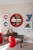 Red plastic child's chair at table on castors, round mirror with red frame between framed pictures on patterned wallpaper