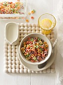 Colourful, fruity cereal rings in a bowl with a jug of milk and a glass of orange juice