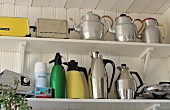 Collection of metal kettles, thermos flasks and retro toasters on white shelves mounted on wooden wall