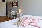 Teddy bear and striped scatter cushions on double bed against grey-painted wall; country-house-style chests of drawers in background