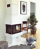 White wood-burning stove and classic chair in renovated, Scandinavian country house with herringbone parquet floor; little boy playing with tablet PC on floor