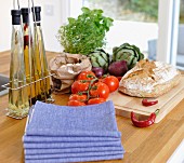Stacked napkins and oil bottles next to fresh vegetables and bread on island counter with pale wooden worksurface