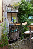 An ornate iron shelf with kitchen utensils and an antique table with garden chairs on a paved area in a garden