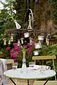 A vase of flowers and tealights hanging from an outdoor chandelier above a bistro table in a garden